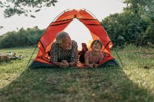 Auf eine Wiese steht ein kleines Zelt. In diesem Zelt liegen ein fröhlicher Opa mit seinem Enkelkind